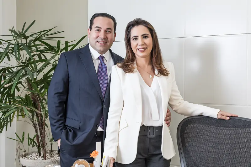 Dois advogados em pé, sorrindo, ao lado de uma cadeira